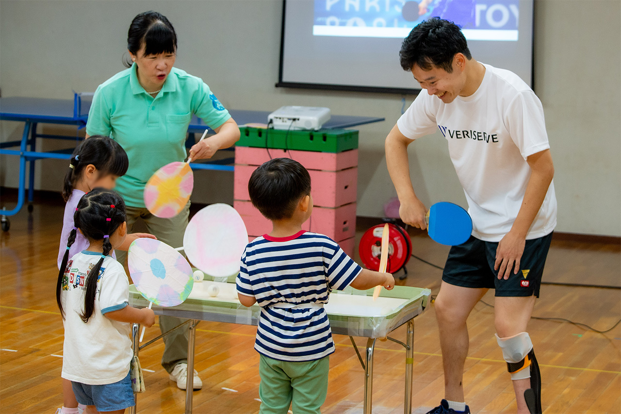岩渕幸洋選手と園長先生がラケットを、子どもたちはうちわを持って、こども園お手製の卓球台で卓球ボールを転がして遊んでいる
